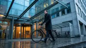Un cadre en manteau conduit son vélo à la main devant l'entrée vitrée d'un immeuble de bureaux un matin d'automne, un abri à vélos en arrière-plan