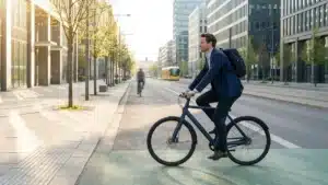 Salarie en veste de costume roulant a velo sur une piste cyclable le long d'un boulevard d'immeubles de bureaux, un matin ensoleille